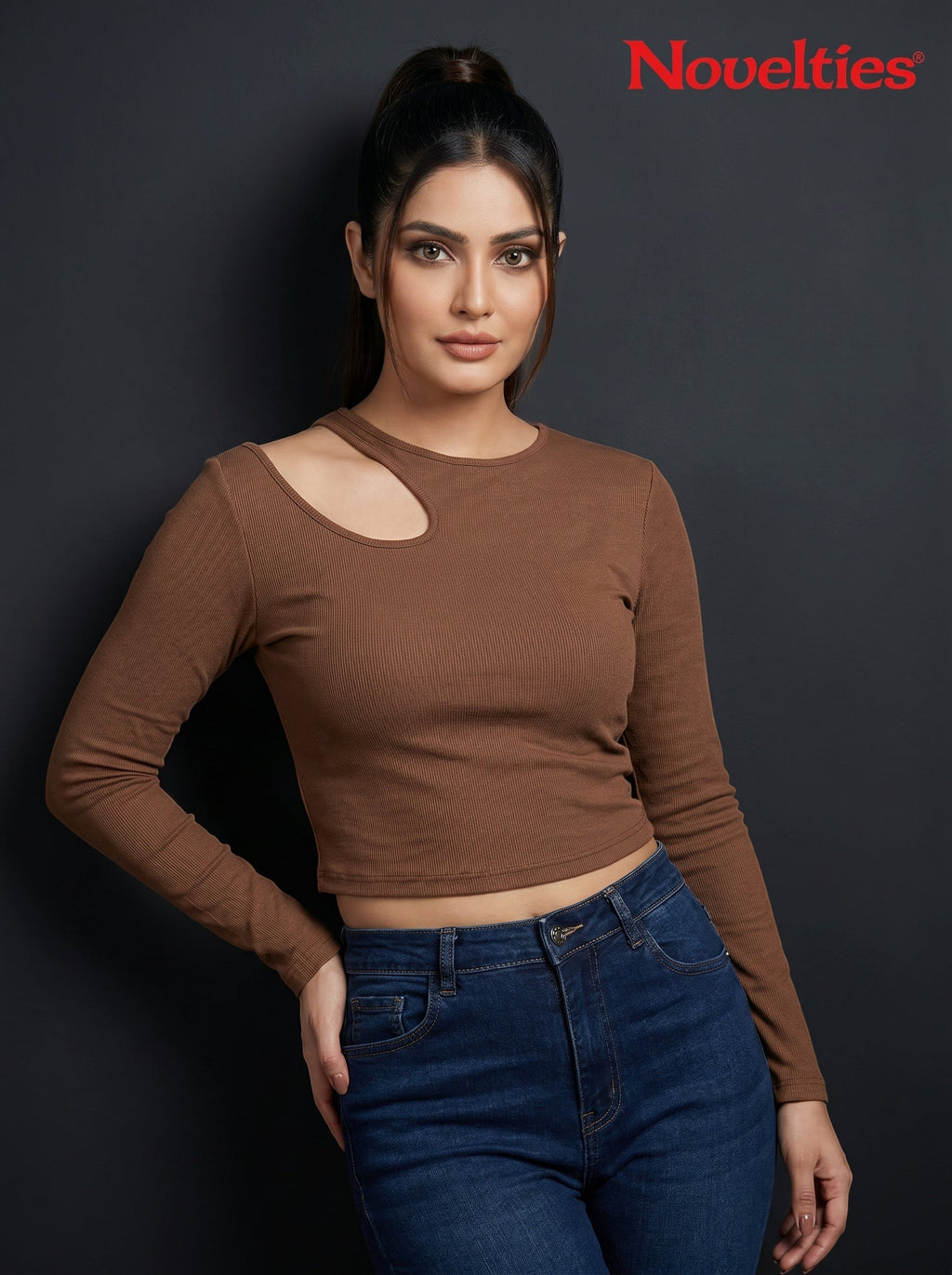 Model wearing a brown Novelties® ribbed long sleeve cropped top with an asymmetric shoulder cutout and crew neck.