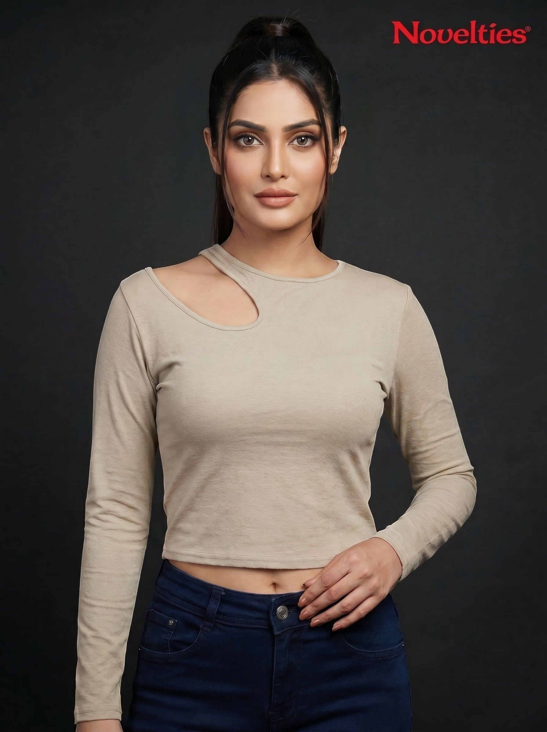 Model wearing a beige Novelties® ribbed long-sleeve cropped top with an asymmetric shoulder cutout against black.