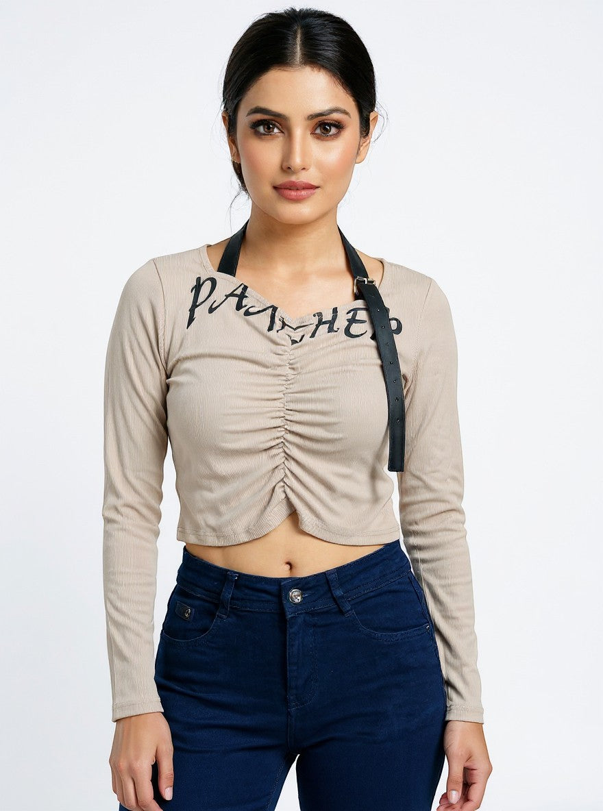 Model wearing a beige Novelties® ruched long-sleeve crop top with graphic script and detachable black strap detail.