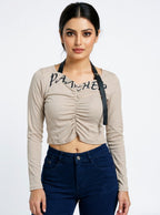 Model wearing a beige Novelties® ruched long-sleeve crop top with graphic script and detachable black strap detail.