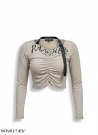 Novelties® beige ribbed long-sleeve crop top with front ruching, black graphic script, and detachable strap detail.