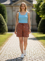 Model wearing Novelties® rust floral print shorts with a blue tank top and white sneakers on a stone path.