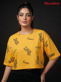 Model wearing a yellow Novelties® oversized crop tee with a playful teddy bear and slogan print on black background.
