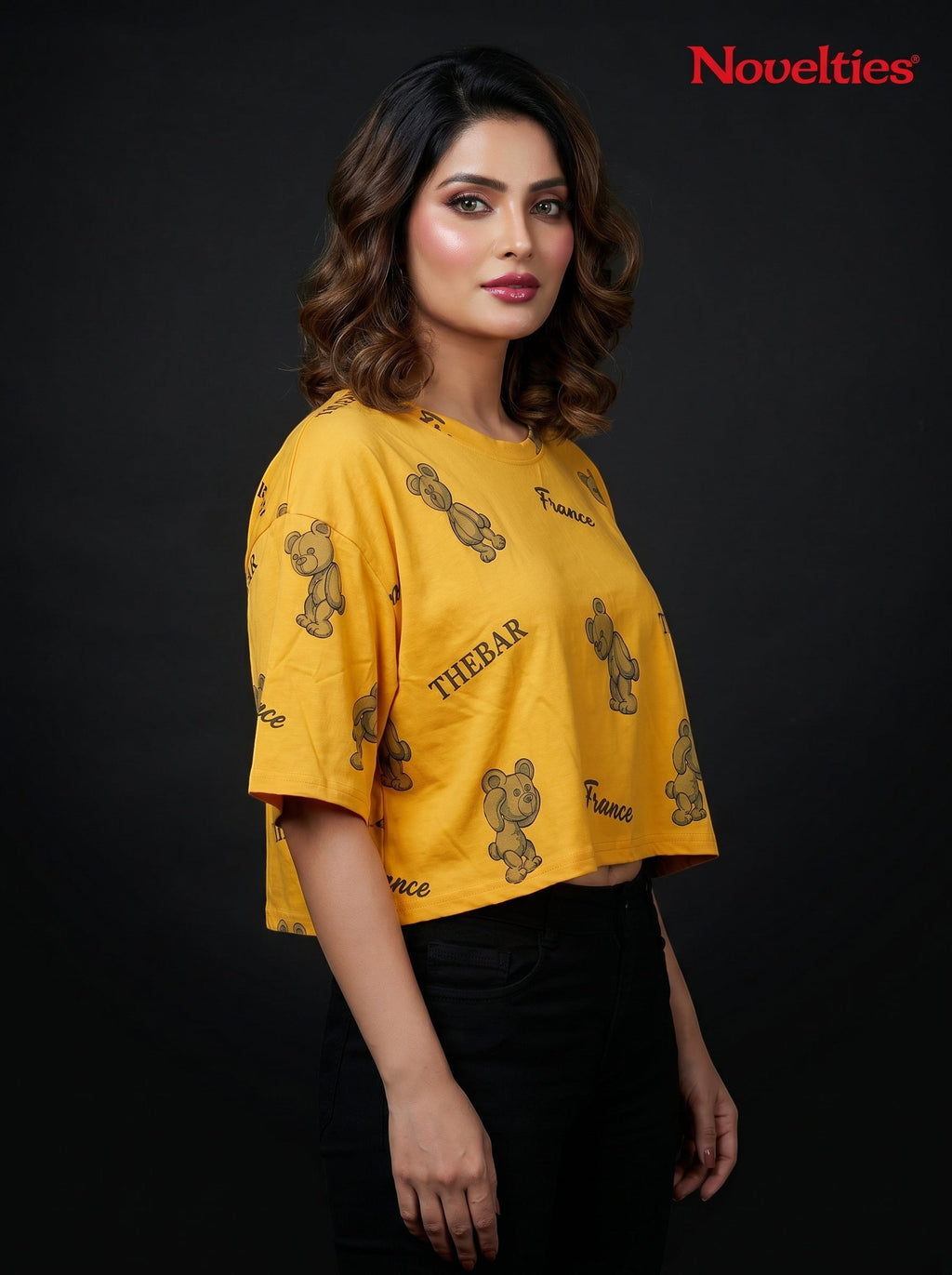 Model wearing a yellow Novelties® oversized crop tee with a playful teddy bear and slogan print on a black background.