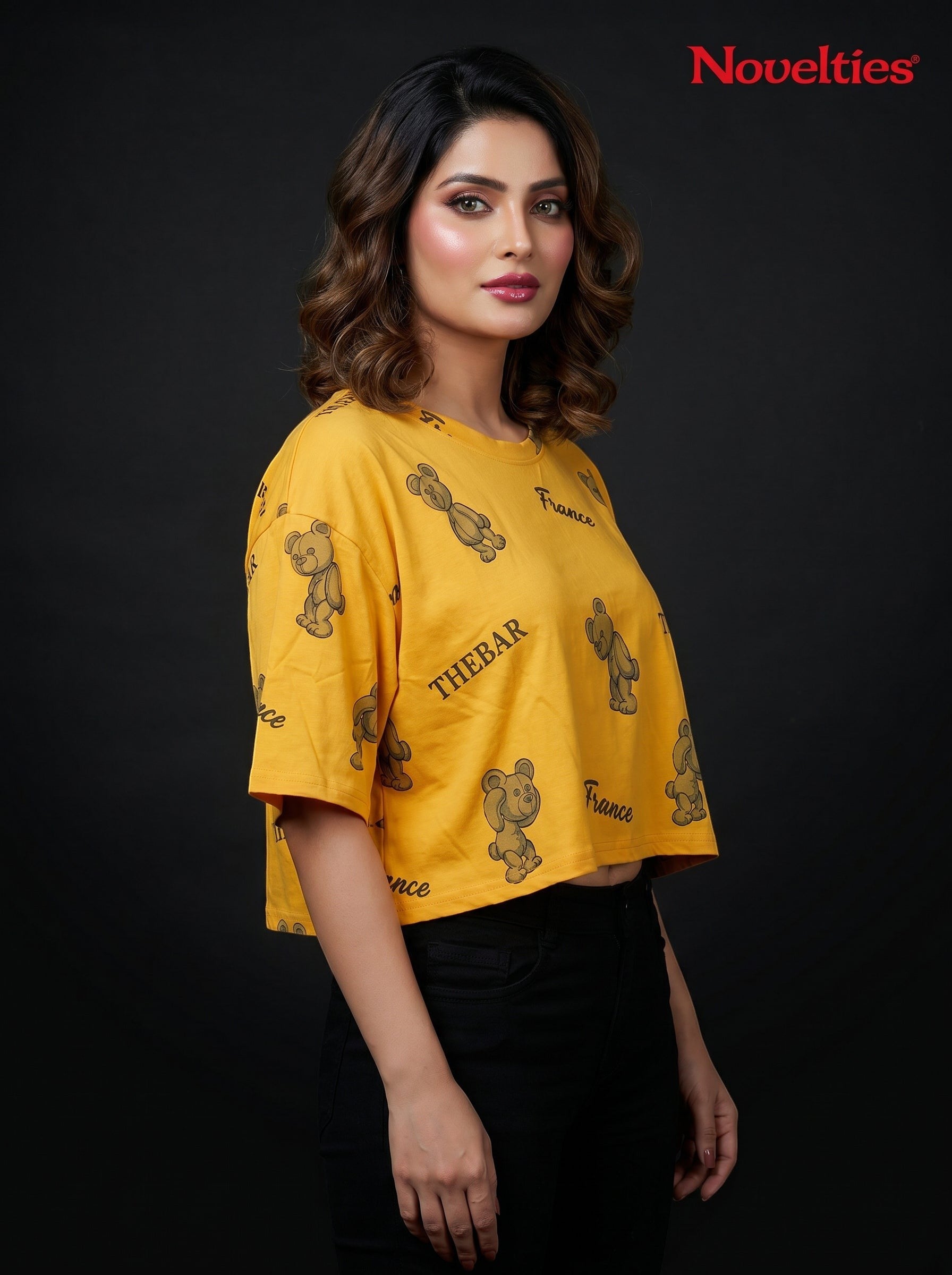 Model wearing a yellow Novelties® oversized crop tee with a playful teddy bear and slogan print on a black background.