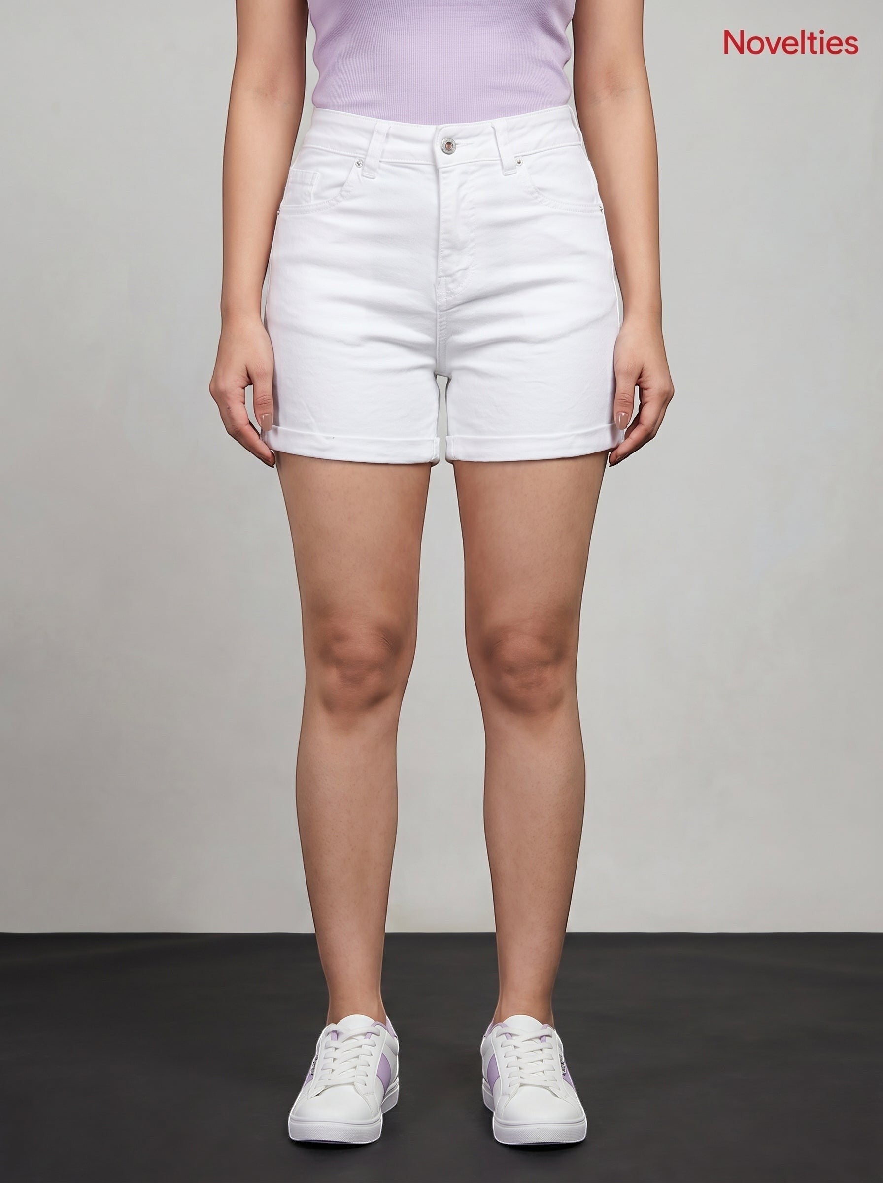 Model wearing Novelties® high-rise white denim shorts with a cuffed hem, paired with a lavender top and white sneakers.